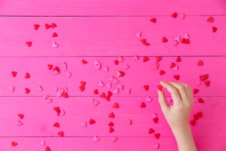 Scattered romantic red hearts symbolising love on a bright exotic pink background with the hand of a young girl in the corner holding one as a Mothers Day, Valentines or anniversary backgroundの写真素材
