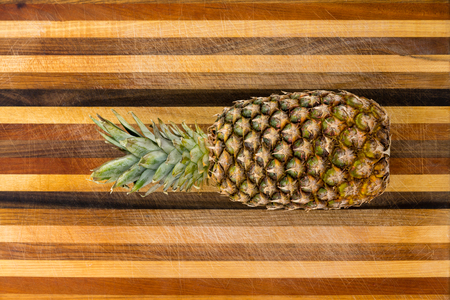Healthy ripe whole pineapple with leaves lying on a decorative bread board with inlaid stripes of wood centered in the middle viewed from aboveの写真素材