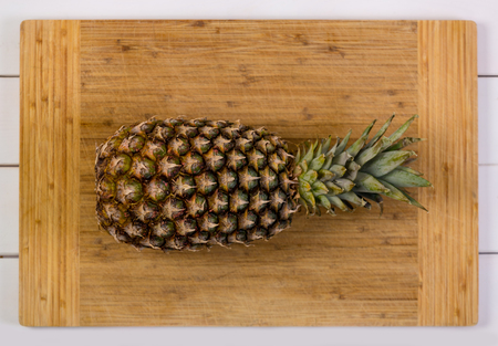 Whole ripe pineapple with leaves on a wooden chopping board over a rustic white wood table viewed from overhead close upの写真素材