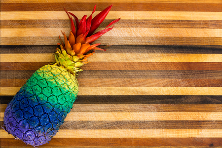 Ripe tropical pineapple with its leaves painted in the colors of the rainbow lying to the side on an ornamental cutting board with inlaid wood stripes and copy space in a close up overhead viewの写真素材