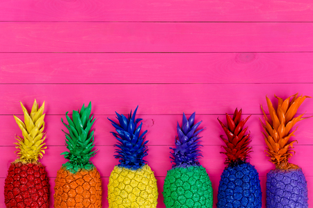 Row of colorful painted pineapples with their leaves in the colors of the rainbow or spectrum on a vivid pink wooden background with copy spaceの写真素材