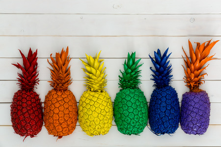 Row of colorful vivid rainbow colored pineapples with their leaves forming a lower border on white wood with copy spaceの写真素材