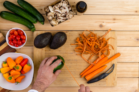 Chef slicing fresh vegetables for cooking on a wooden chopping board holding a green pepper with peeled carrots, zucchini, mushrooms, tomatoes,ripe avocado pears and carrots in an overhead viewの写真素材