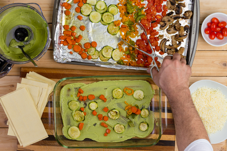 Male chef preparing a vegetable lasagne layering mashed avocado pear, pasta and diced fresh vegetables with a pair of tongsの写真素材