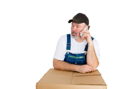 Workman in overalls listening to a phone call on his mobile phone with a serious expression as he leans on a brown cardboard box isolated on whiteの写真素材