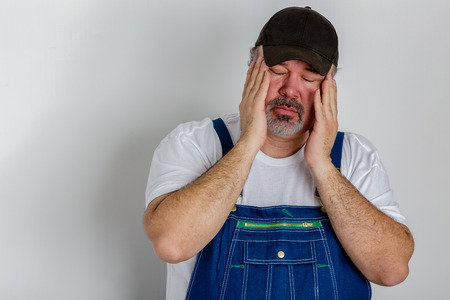 Worker in overalls with a bad headache holding his head in his hands with his eyes closed against the painful throbbing isolated on whiteの写真素材