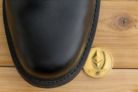 Single Ethereum cryptocurrency coin under a black leather shoe on wooden floorboards viewed from above in a conceptual imageの写真素材