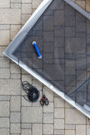 Replacing damaged wire mesh on a patio screen door in an overhead view of the partially finished metal frame, molding and scissors on a brick patioの写真素材