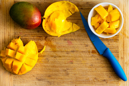 Sliced ripe mango in a bowl and hedgehog shape on a bamboo cutting board with colorful blue kitchen knife and copy spaceの写真素材