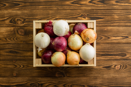 Assorted colors of fresh whole onions in a small wooden box displayed on a rustic table wooden table at an organic farmers market viewed close up from above in a healthy diet conceptの写真素材