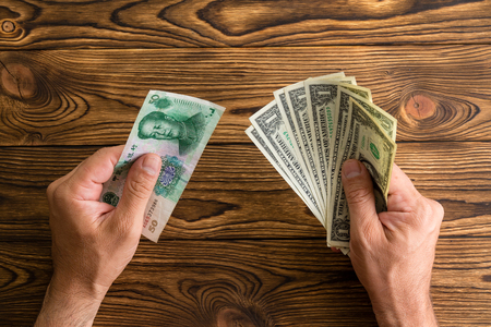Man holding Chinese Yuan and a fanned fistful of United States Dollars in his hands over a wooden table viewed from above in a monetary foreign exchange and currencies conceptの写真素材