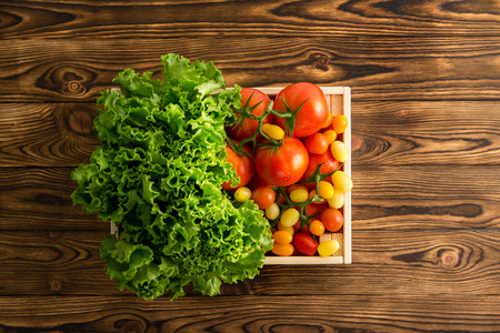 Fresh head of leafy green lettuce and a variety of ripe juicy tomatoes in a small wooden crate at wooden table at an organic farmers market on a wooden table viewed top downの写真素材