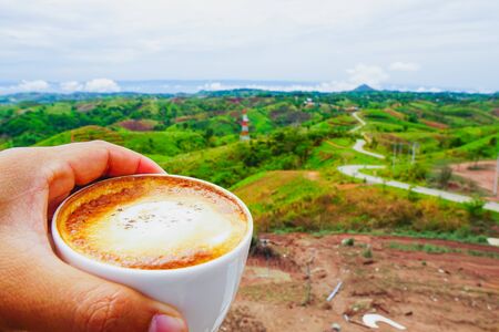 The hand hold the white hot coffee mugs with green fresh mountain and morning fog as background.の写真素材