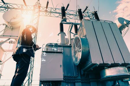 Businessman wearing a black suit, standing looking at a large power transformer with orange sunlight...