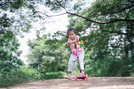 Asia children learn to ride scooters in a park on a summer day. Preschooler girl riding a roller. Kids play outdoors with scooters. Active leisure and outdoor sport for children.の写真素材