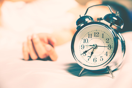 Young hates waking up early in the morning. Sleepy girl looking at alarm clock and trying to hide under the pillowの写真素材