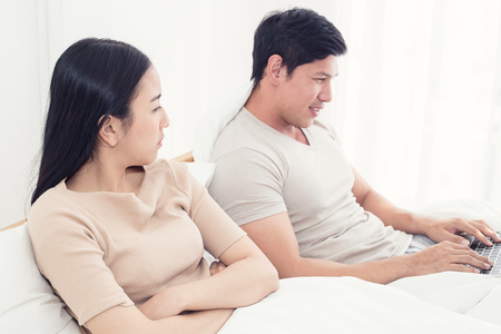 Couple in bed, happy smiling man turned her back to man, reading message on phone from her lover, worried woman lying next to her, trying to peek at screen. Cheating and infidelity conceptの写真素材