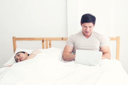 Couple in bed, happy smiling man turned her back to man, reading message on phone from her lover, worried woman lying next to her, trying to peek at screen. Cheating and infidelity conceptの写真素材