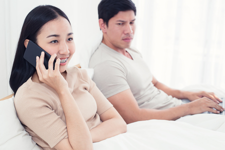 Couple in bed, happy smiling man turned her back to man, reading message on phone from her lover, worried woman lying next to her, trying to peek at screen. Cheating and infidelity conceptの写真素材