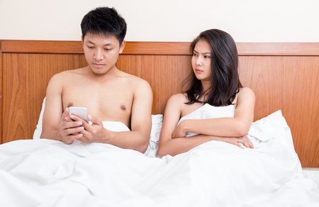 Couple in bed, happy smiling man turned her back to man, reading message on phone from her lover, worried woman lying next to her, trying to peek at screen. Cheating and infidelity conceptの写真素材