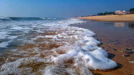 A tranquil beach scene featuring gentle waves lapping against the sandy shore. The foamy surf and clear blue sky create a serene atmosphere perfect for relaxation and escape.の素材