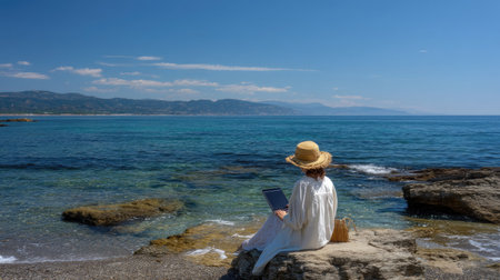 A woman sits on a rocky beach, working on her laptop under a bright sky. The tranquil waters and stunning coastline create a perfect setting for remote work and leisure.の素材