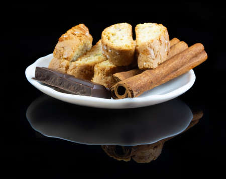 Cinnanon sticks, chocolate and bisquits in a white plate over black reflective backgroundの写真素材