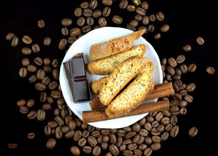 Cinnanon sticks, chocolate and bisquits in a white plate surrounded by coffee beans over black reflective backgroundの写真素材
