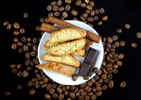 Cinnanon sticks, chocolate and bisquits in a white plate surrounded by coffee beans over black...