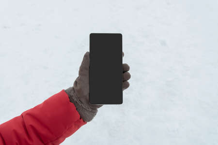 Female hand in winter gloves holding smart phone with blank screen for copy space, close up, snow...