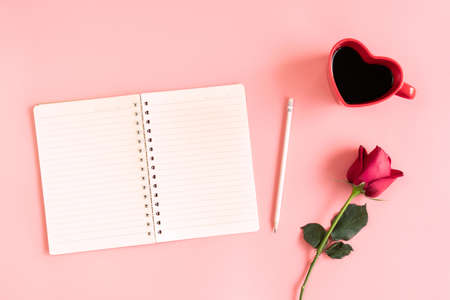 Valentine's Day background. Heart shaped cup of black coffee, red rose and notebook on pastel pink...