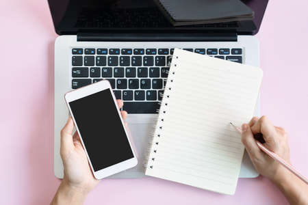 Flat lay of woman hand writing on notepad with laptop on pink table. Business and technology. family expense summary concept. Top view and copy space.の写真素材