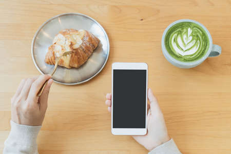 Flat lay of women's hands using mobile phone while eating croissant with a cup of green tea latte on wooden table in cafe. Food and beverage, technology and lifestyle concept. copy space, top viewの写真素材