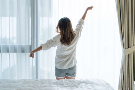 Back view of beautiful young Asian woman in white pajamas stretching after wake up in the morning in bedroom.の写真素材