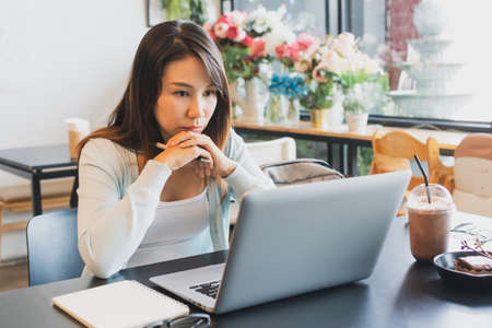 Stress Asian woman freelancer working on laptop with notebook, glasses, drinks and cake on table in coffee shop. Business, lifestyle and technology concept. Close upの写真素材
