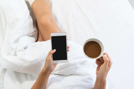 Woman relax on the bed using mobile phone and cup of coffee in hands, top view and copy space.の写真素材