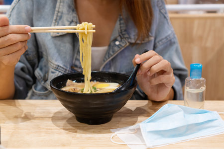 Young Asian girl eating ramen with sanitizer gel and surgical face mask on wooden table in restaurant. New normal lifestyle, health care concept. Close upの写真素材
