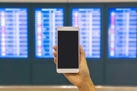 Hand of young Asian tourist woman holding smartphone near flight information board in international airport. Close up, copy spaceの写真素材