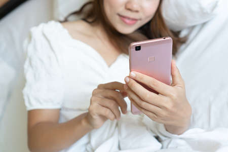 Beautiful young Asian smiling woman lying in white bed and using a phone in her bedroom. Technology, free time, communication concept. Close upの写真素材