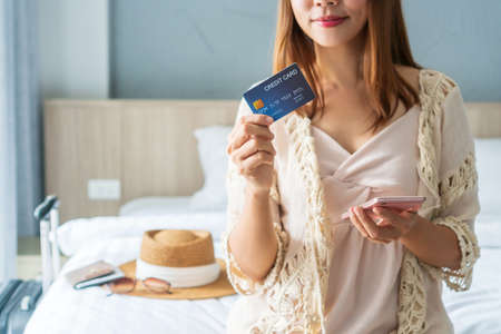 Happy traveler Asian woman holding credit card while using smartphone with travel accessories on bed. Online shopping and travel, cashless society, technology and lifestyle conceptの写真素材