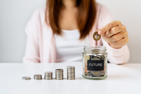 Hand of Asian woman putting coin in glass jar on white table background. Saving, collect money for future, investment concept. Close upの写真素材