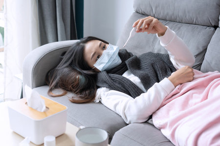 Young Asian woman get sick with changing seasons lie down on couch. Girl feel cold and has a high fever lying on sofa in living room at home. Health problems, cold and flu concept. Closeupの写真素材