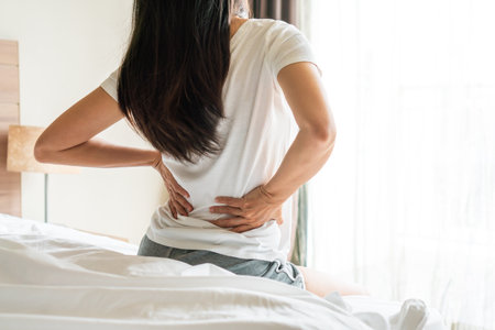 Back view of Asian woman suffering from backache sitting on bed in bedroom at home. Unhappy girl feel back pain due to mattress too soft. Health problem concept. close up, copy spaceの写真素材