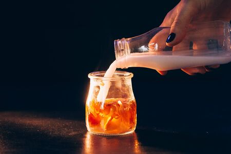 Girl pours milk from a glass bottle into a glass jar of tea on a black background with warm light