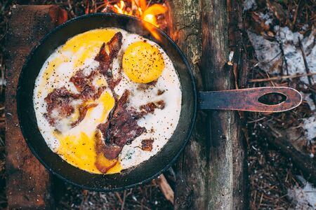 Fried eggs with bacon in a pan in the forest. Food at the camp. Fried egg with bacon on fire. Picnicの写真素材