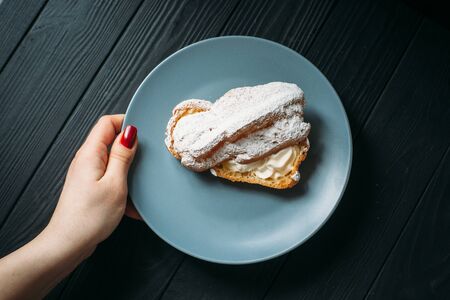 Hand holding a plate with eclair. Eclair with half-cream under powdered sugar.