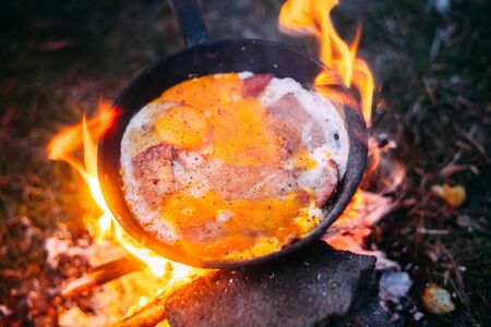 Fried eggs with bacon in a pan in the forest. Food at the camp. Scrambled eggs with bacon on fire. Lots of seasonings on fried eggs. Picnicの写真素材