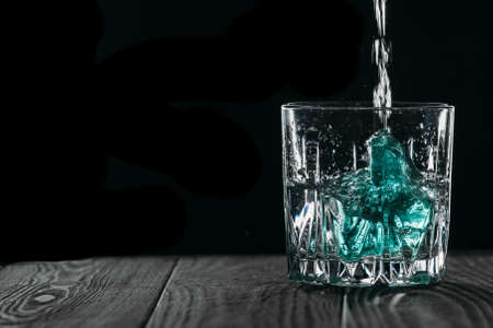 Glass with blue ice cubes. Pouring gin into a glass on a wooden table. Gin in a glass on a black background.の写真素材
