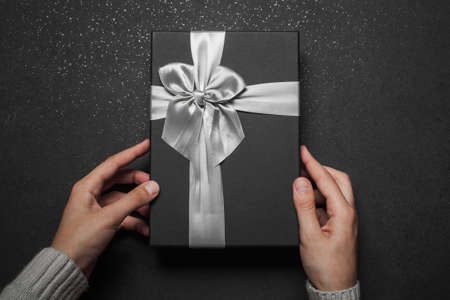 Hands in a sweater opening a black gift box with a silver bow on a black granite background.