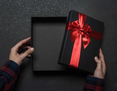 Hands in a shirt open a black gift box with a red bow on a black granite background.